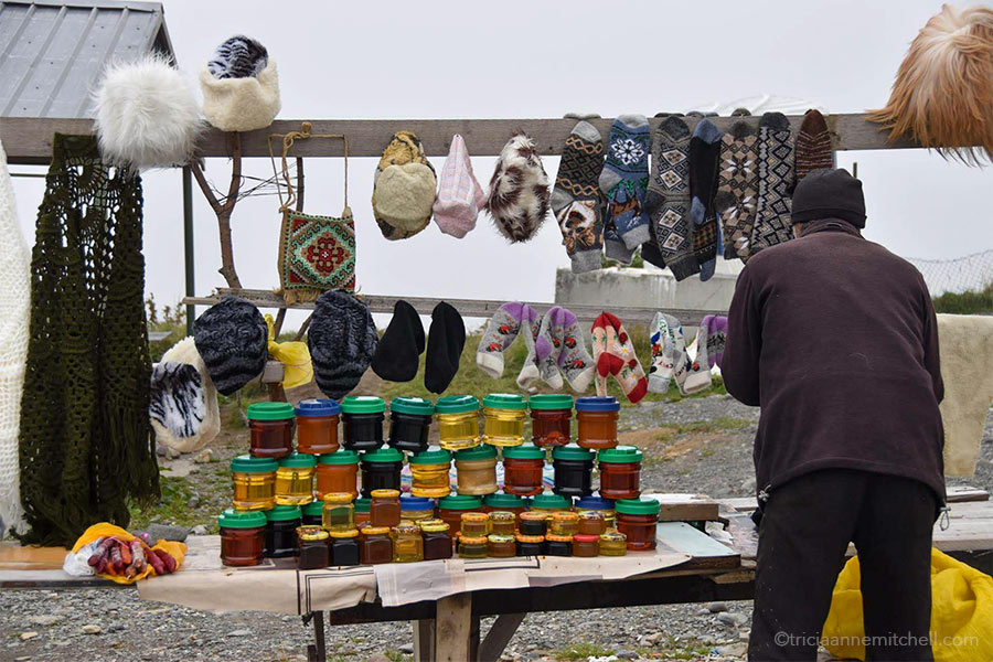 A man sells souvenir items at the Russia–Georgia Friendship Monument. On the table there are 3 rows of jarred honey stacked on one another, along with wool socks, fuzzy hats, and a cross-stitched purse.