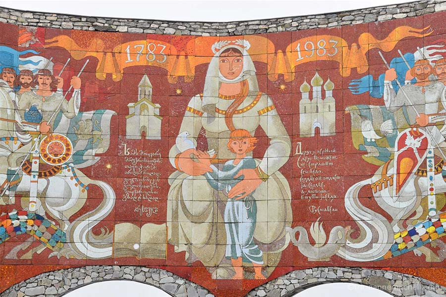 A close-up view of the Russia-Georgia Friendship monument. The background is red, and there is an orange banner that says "1783, 1983." A woman holds a white dove and a child in her hands. Warriors seated on white horses are on either side, as are depictions of two church buildings. 