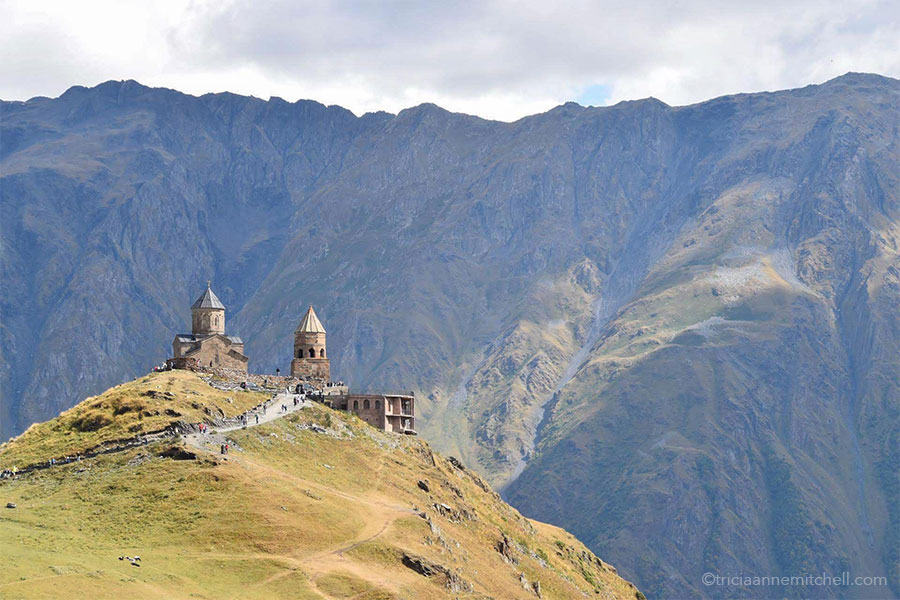 The two rounded towers of the Gergeti Trinity Church rise from a grassy and rocky hilltop. In the background are the steep slopes of the Caucasus Mountains.