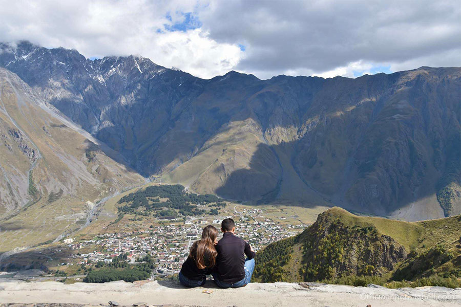 Two people (whose backs are facing the camera) look at the view from the Gergeti Trinity Church. Large mountains are visible off in the distance, and you an see the tiny town of Kazbegi below.