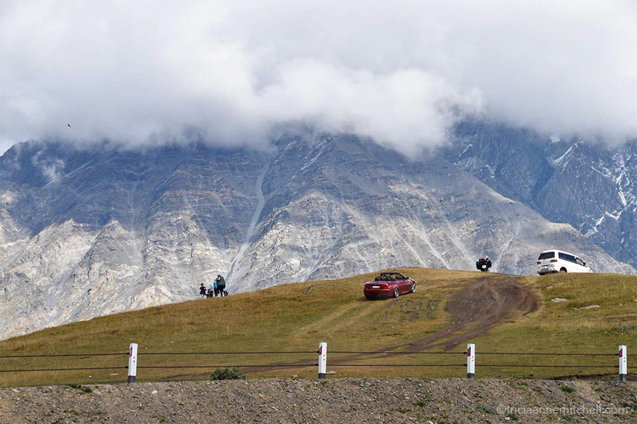 A car and a van drive up the grassy slopes near the Gergeti Trinity Church, marring the landscape. In the background are steep mountain slopes — some covered with a bit of snow.
