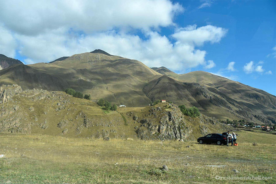 A car parked on a grassy lawn near the Georgian Military Highway. There are greenish-brown mountain slopes in the background. A tiny house sits on one hilltop.