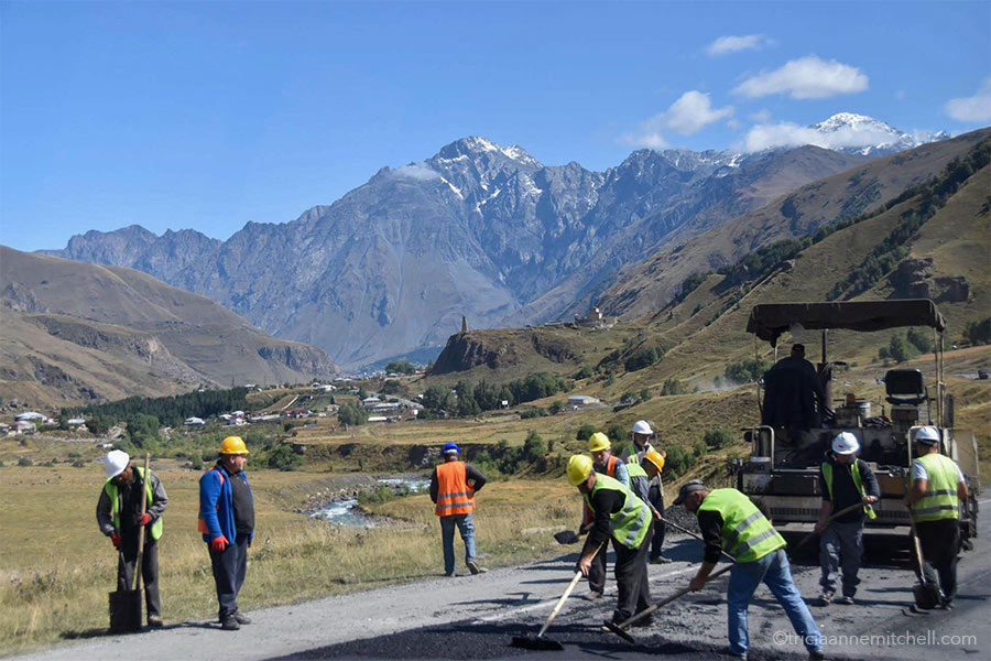 Construction men repair a stretch of road along the Georgian Military Highway. There are snow-capped mountain peaks and a stone watchtower off in the distance.