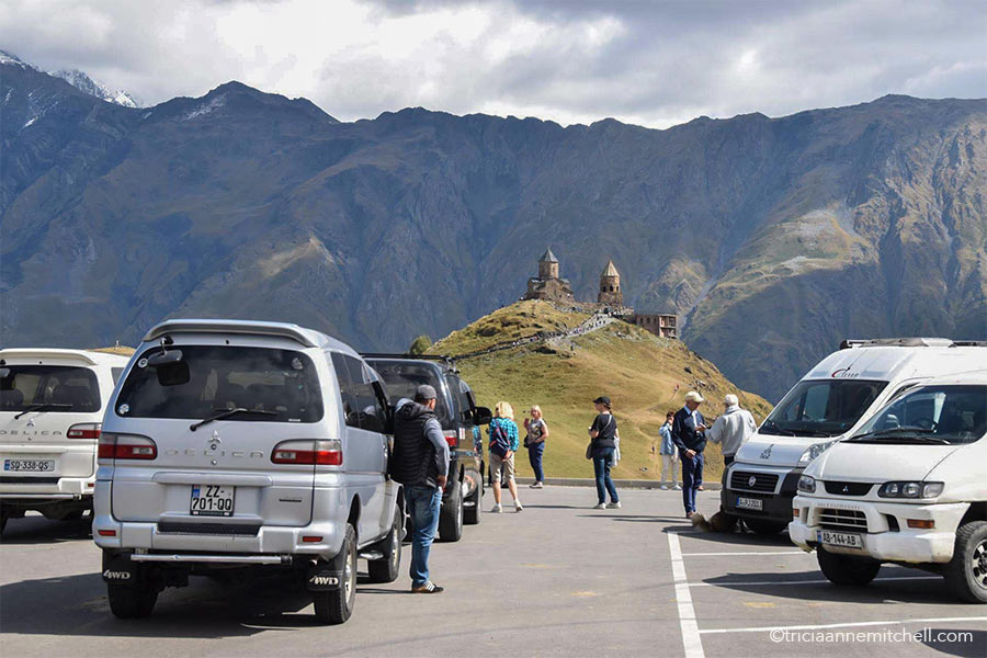Five 4x4 minivans park in a concrete parking lot in front of Georgia's Gergeti Trinity Church.