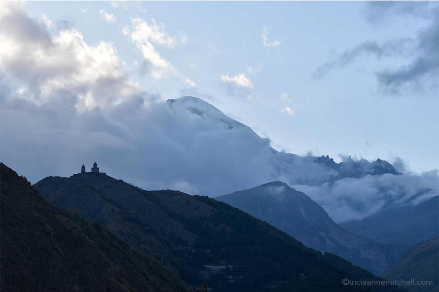 The Gergeti Trinity Church is seen in silhouette form against a cloudy backdrop. Snowy Mount Kazbek is only partially visible.