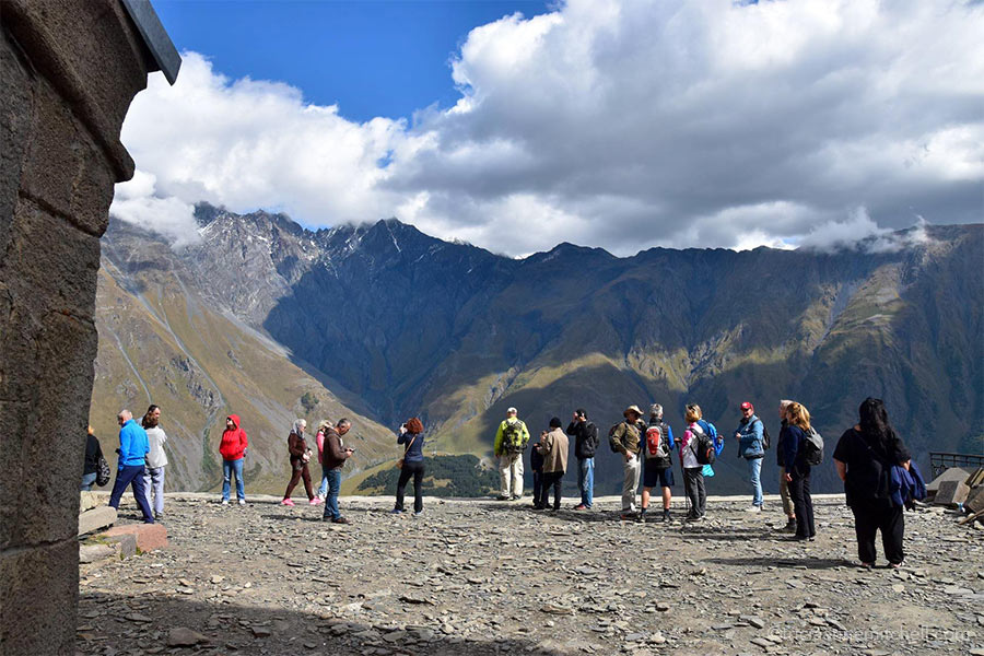 About 15 visitors stand on a rocky platform overlooking a mountainous backdrop near the Gergeti Trinity Church in Georgia. The sky is mostly cloudy.