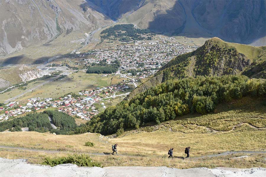 Three hikers walk on a dirt path as they make their way up to the Gergeti Trinity Church. You can see the rooftops of Gergeti and Kazbegi below, and there is a green hilltop.