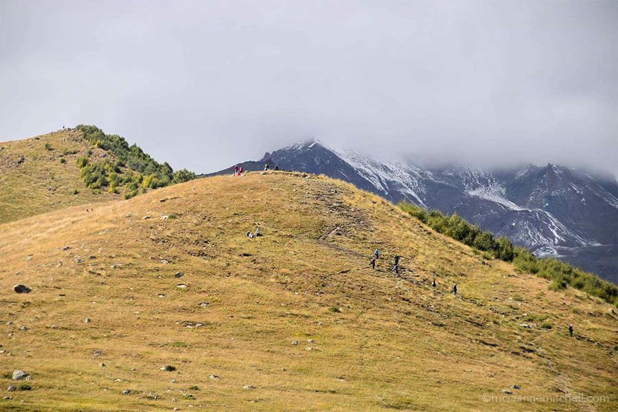 About 10 hikers make their way down a trail. Georgia's Mount Kazbek is concealed by clouds.