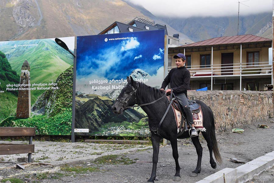 A young boy rides a black horse on a sidewalk in the town of Kazbegi (Stepantsminda), Georgia. There are 2 tourism billboards on his right, and a house in the background.