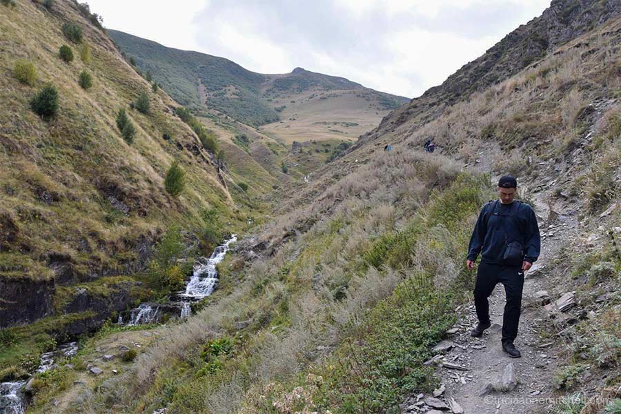 A man hikes down a rocky/dirt path alongside a stream in the Caucasus Mountains.