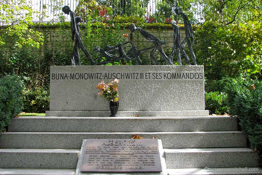 A rectangular headstone features abstract concentration camp figures. Inscribed on it are the words "Buna Monowitz Auschwitz III et ses kommandos." Below another plaque reads, "Auschwitz, n'oublions jamais."