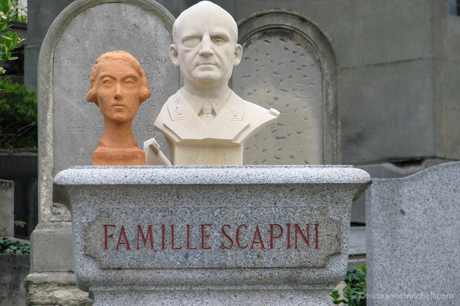 Two busts - one female and orangish, and another male and white - stand atop a headstone. The words "Famille Scapini" are written on it.