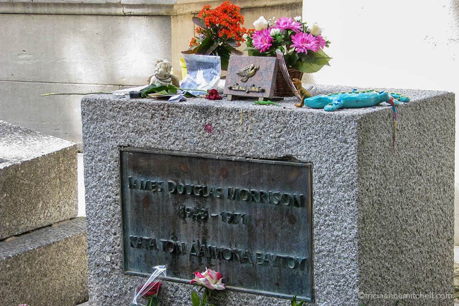 Jim Morrison's headstone in Paris' Pere Lachaise Cemetery. There are several items sitting on top of the square stone memorial including an angel's head, flowers, and a green clay lizard. The headstone reads: "James Douglas Morrison, 1943-1971"