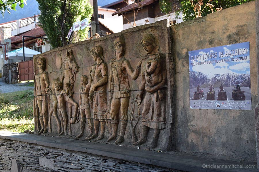 Above a bench in Kazbegi, Georgi the wall is decorated with folk art figures, including adults, children, and a horse. A poster for Kazbegi quad tours also adorns the wall.