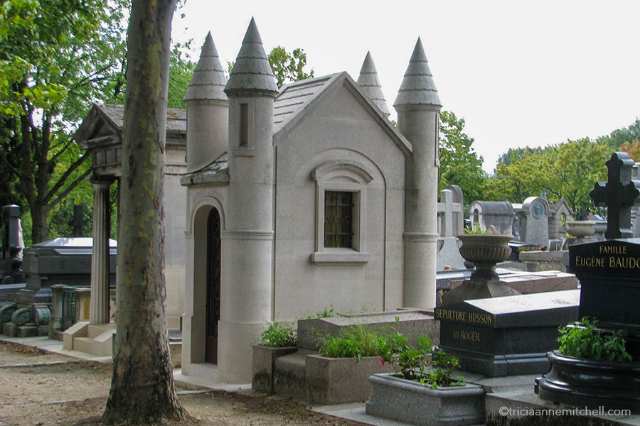 A stone mausoleum resembling a small castle with four towers sits beside other headstones in the Pere Lachaise Cemetery.