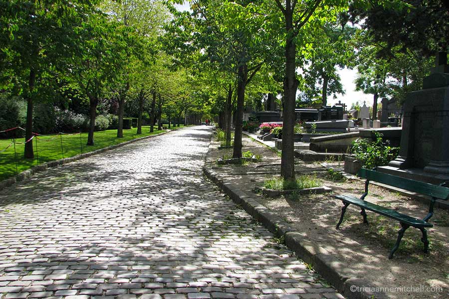 A brick street in Pere Lachaise Cemetery is partially cast with shadows from green trees overhead. To the right there is a green bench. Headstones are visible in the background.