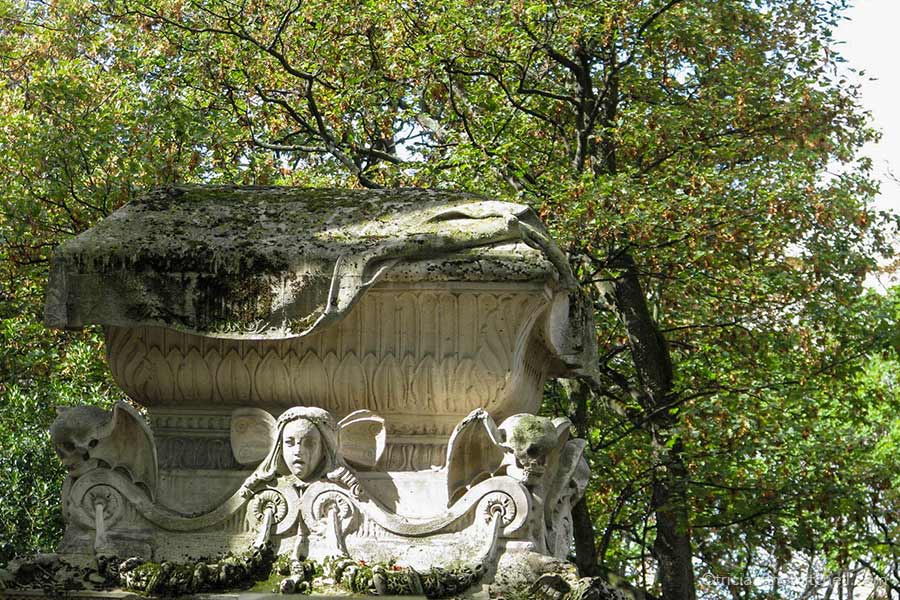 Close-up of a stone mausoleum decorated with a human head flanked by two skulls with wings. There is a tree overhead.