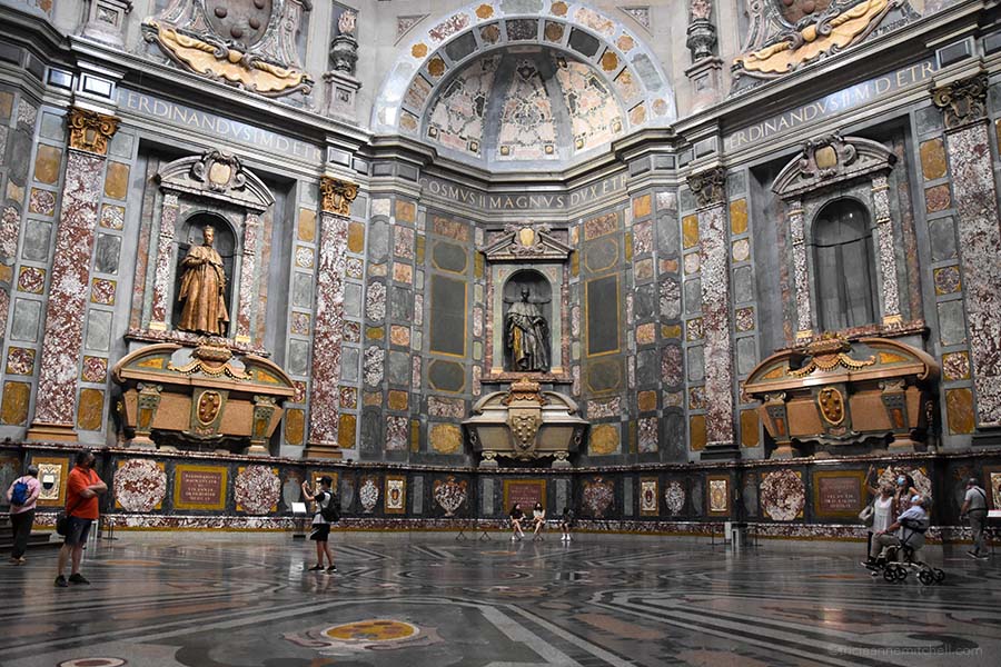 The interior of the Chapel of Princes, in the Medici Chapel in Florence, Italy. Here, several tourists look at the ornately decorated walls as well as the stone sarcophagi.
