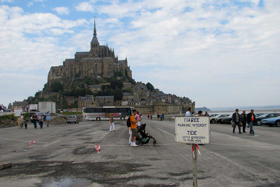 Mont Saint Michel's parking lot in 2011. Tourists, cars, and a bus are in the lot. There is a sign to say that parking in one section is forbidden.