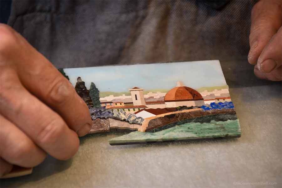The master artisan assembles individual fragments of stone in order to complete a Florentine mosaic that depicts the Duomo, trees, and other towers in Florence, Italy.
