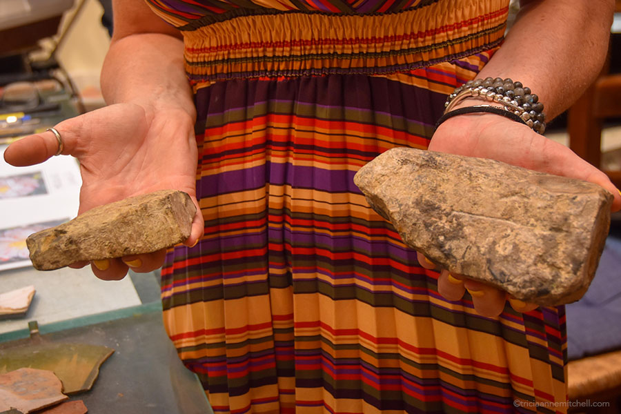 A woman in an artisan's workshop holds two rocks in her hand as she explains the process of making Florentine mosaics / Florentine marquetry.