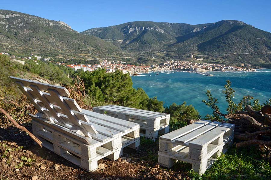 White wooden pallets that have been turned into furniture sit on a hillside overlooking a mountainous, coastal view