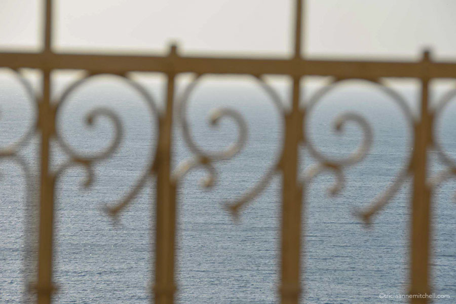 The swirly embellishments of an iron railing, with the blue sea in the background
