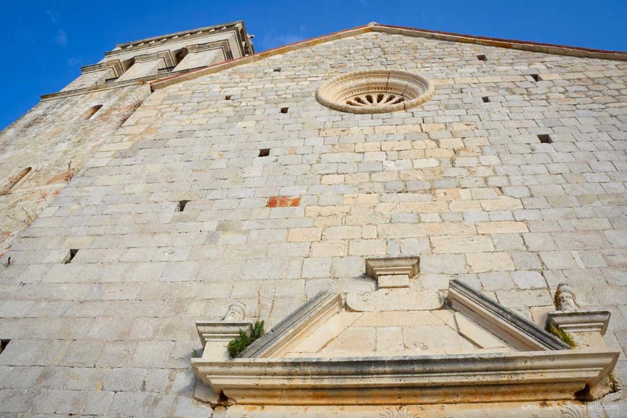 Close-up detail of a white stone church in Croatia. Above the doorway, there are 2 figures of male heads with beards.