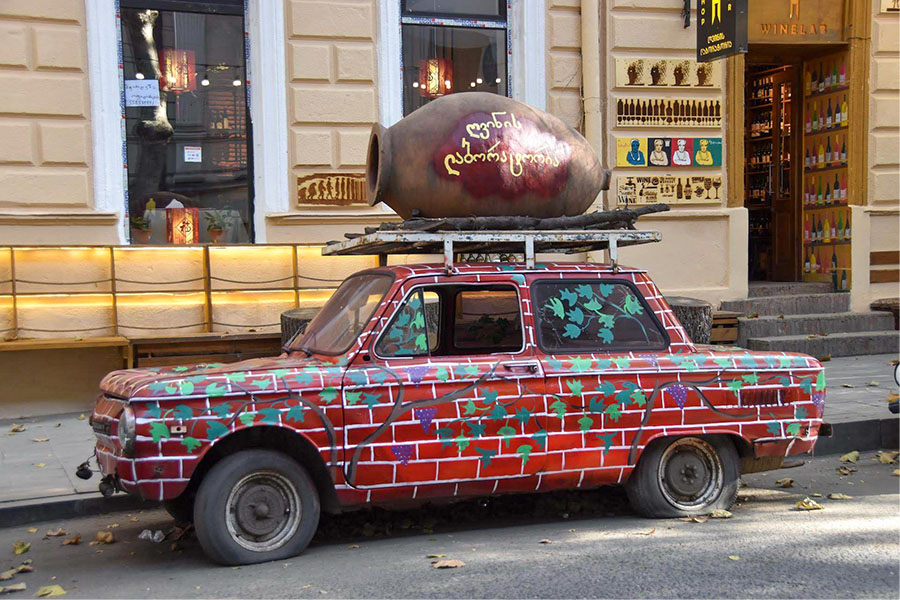 A qvevri sits atop a red vintage car in front of a wine establishment in Tbilisi, Georgia.