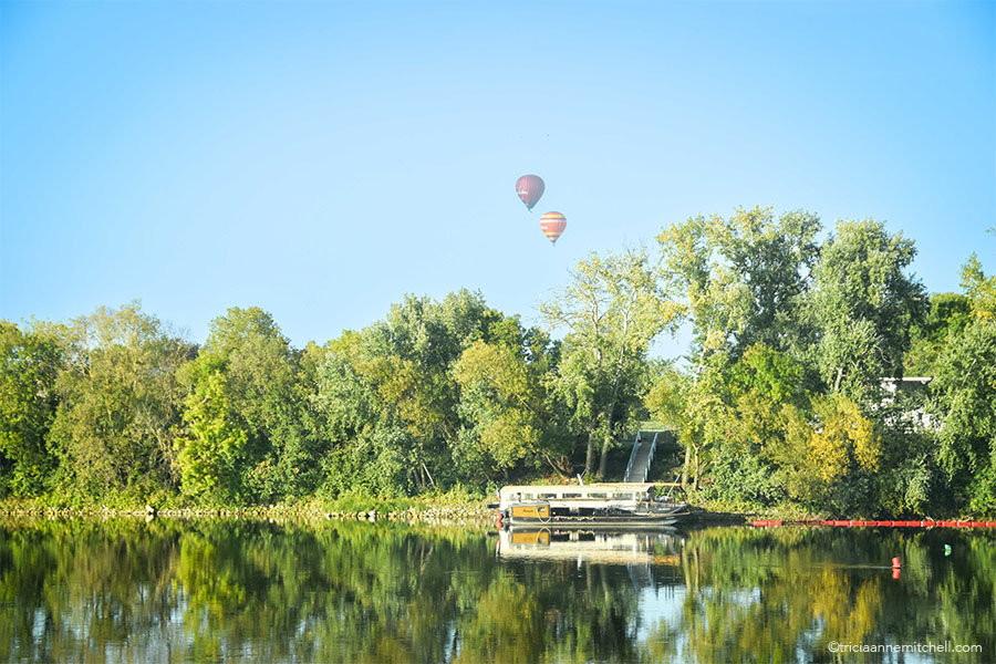 Two hot air balloons fly over a forested island near the Loire River in France. A boat is tied up on the shore.