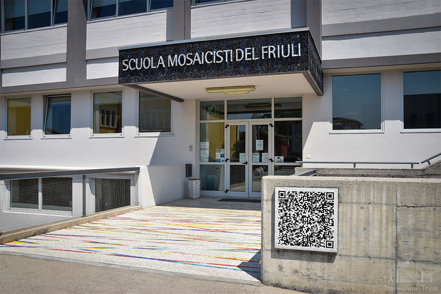 The modern entrance to Spilimbergo's Mosaic School. Above the doorway is an overhang with the words "Scuola Mosaicisti del Friuli" written on it. A mosaic QR code panel hangs on the concrete wall.