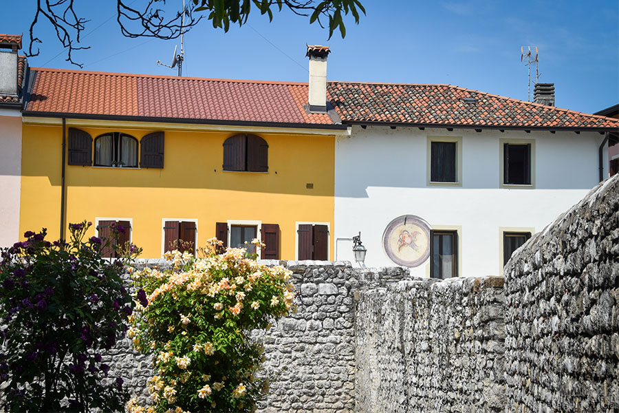 Two adjoining homes in Spilimbergo, one yellow-orange, the other white. Both buildings have terracotta tiles and stone walls.