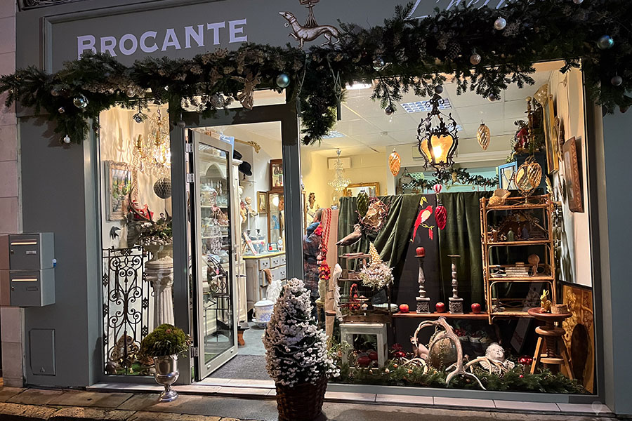 The storefront of the Brocante l'Hermine is decorated for Christmas with green fabric, a snow-covered evergreen tree, and a red parrot and ornaments.
