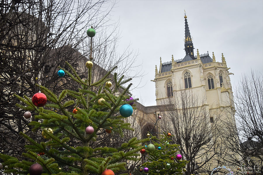 Amboise's Saint Hubert Chapel towers over the Michel Debré Square on an overcast day. Green evergreen trees, decorated with multicolored ornaments, frame the scene.