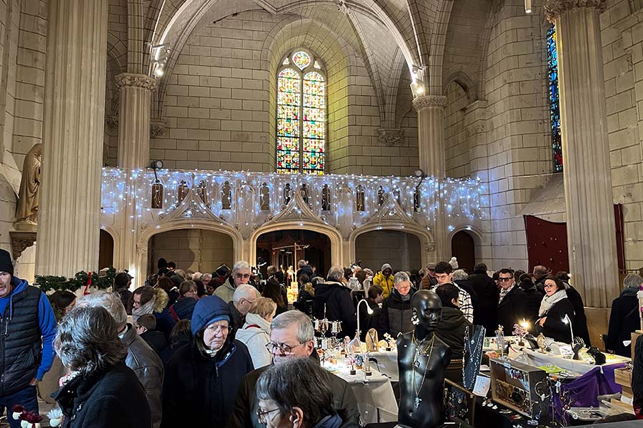The interior of Amboise's Saint Florentin Church is decorated with pale blue Christmas lights below a colorful stained glass panel. Below, Christmas market attendees peruse tables filled with jewelry for sale.