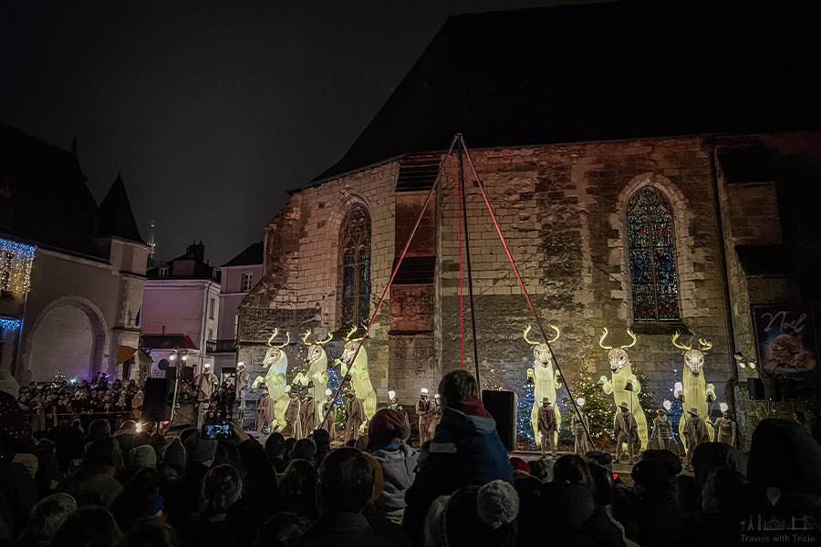 Six oversized glowing reindeer puppets stand in a winter holiday performance in Amboise, France alongside the stone exterior of Saint Florentin Church. Spectators watch the spectacle, with some children sitting atop the shoulders of adults.