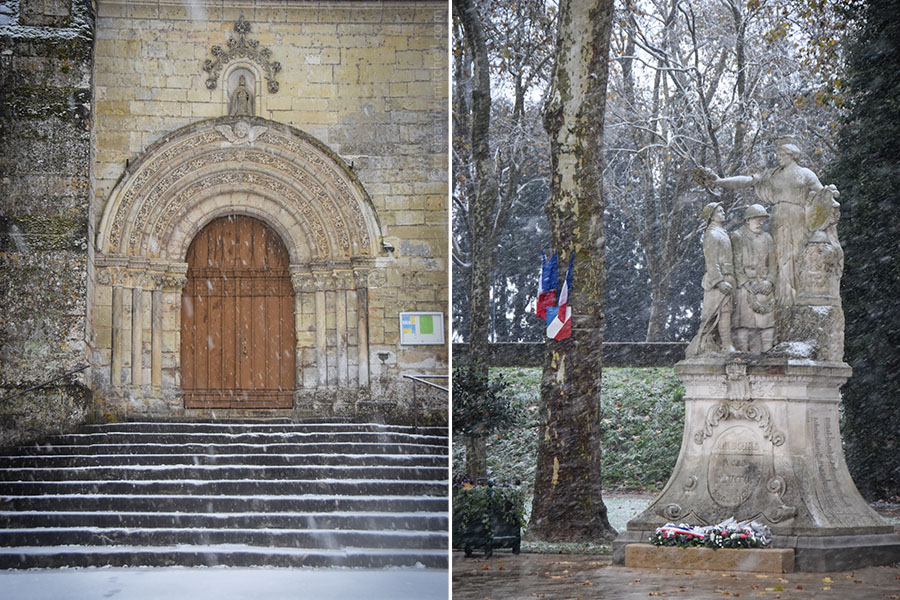 On the left, the main entrance of Saint Denis Church in Amboise, France is visible. There is snow on the stairs leading into the church. On the right, Amboise's War Memorial is lightly dusted in snow. There are three figures in the memorial. Several French flags adorn a Plane tree to the left, and bouquets of flowers sit at the foot of the memorial.