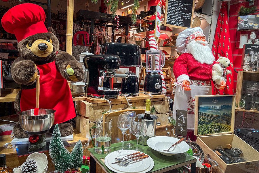 The "Un Coin de France" shop in Amboise, France is seen with Christmas decorations and products for sale in the window. There are green and red dishes, drinking glasses, and stuffed animals, including a bear and Santa Claus wearing a chef's hat.