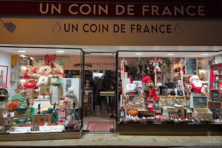 The "Un Coin de France" shop in Amboise, France is seen with Christmas decorations and products for sale in the window at night. There are green and red dishes, drinking glasses, and stuffed animals, including a bear and Santa Claus wearing a chef's hat.