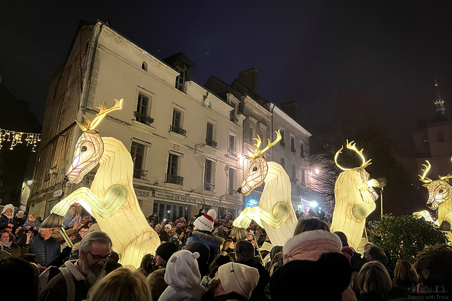 Four glowing reindeer puppets move through the main street of Amboise, France during a holiday parade. Crowds line both sides of the street.