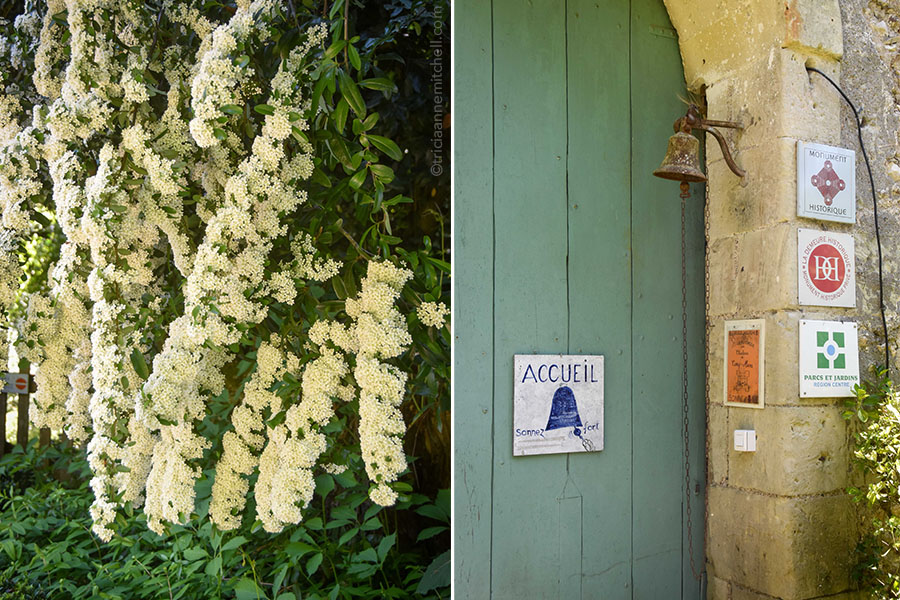 On the left, branches filled with white bridalwreath flowers hang in the courtyard of the castle in Cinq Mars la Pile, France. On the right, a bell hangs on a stone archway, near a green wooden door. Signs posted on the door, written in French, indicate that this is a historic monument and that visitors to the castle should ring the bell forcefully.