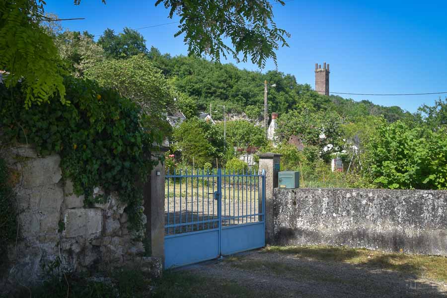 The Pile de Cinq Mars (a brick Gallo-Roman tower) rises above the treetops on a hillside in the town of Cinq Mars la Pile, France. In the foreground is the entryway to a home with a blue gate.