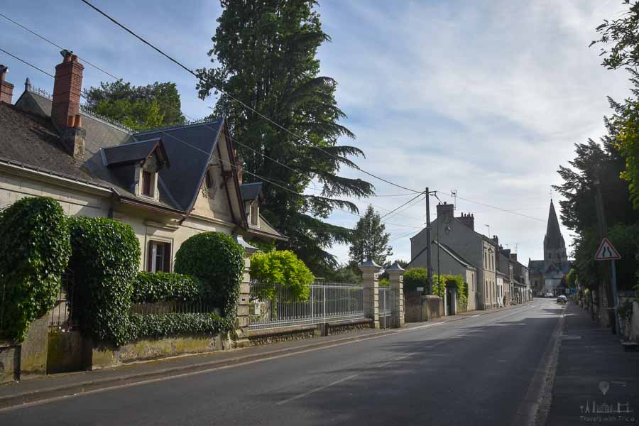 The two-lane road heading into the town of Cinq Mars la Pile, France. The town's church steeple is visible at the end of the road, and an old home is on the left side of the road.